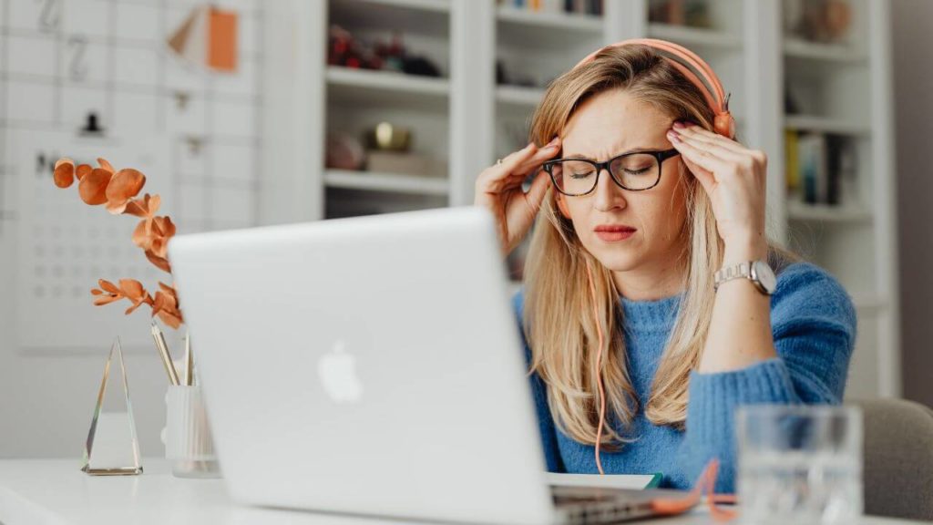 Woman sitting at a desk behind a laptop holding her head as if she is experiencing a headache